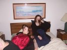 Hawk and Aisling and Kendra wearing red satin pajamas drink coffee in bed beneath an ulgy hotel watercolor.  Photo also features Jean-Claude's ear